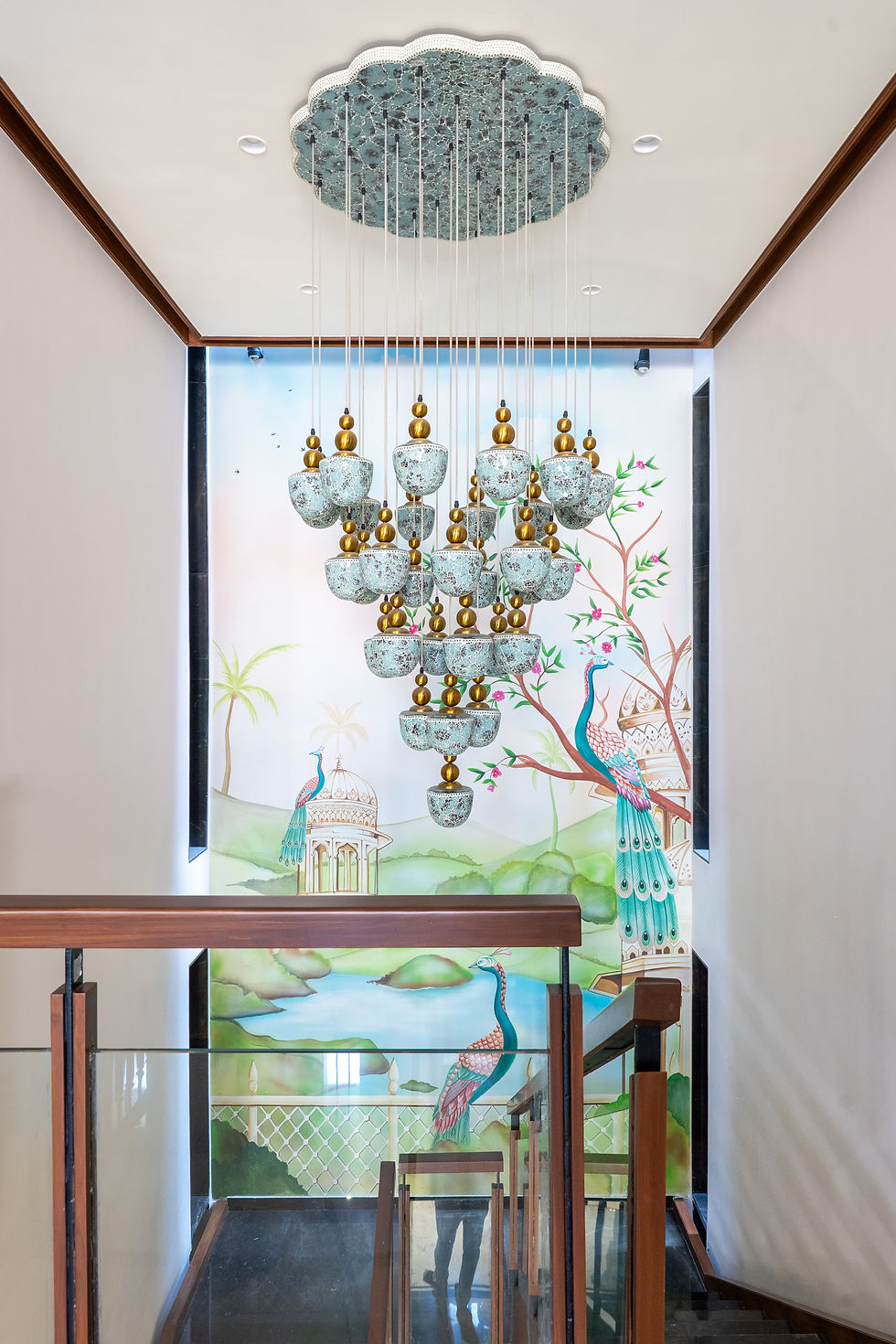 Elegant stairwell with a hanging blue-and-gold chandelier above a peacock mural and glass-railed stairs.