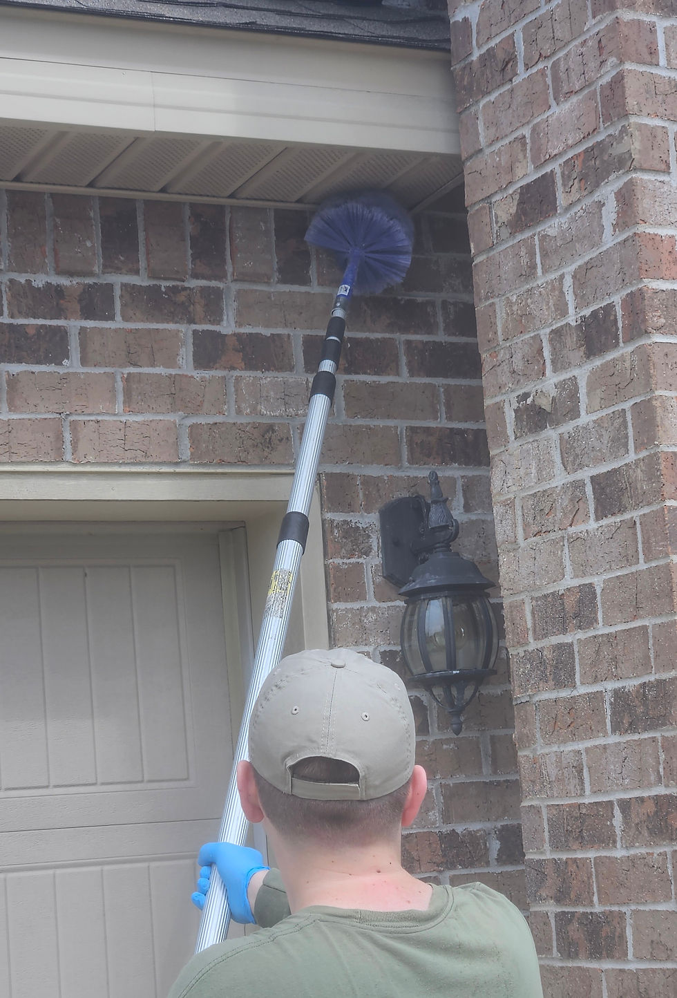 Man in cap and mask pries open window, wearing gloves, outside a house with green ivy. Bright daylight creates tense mood.