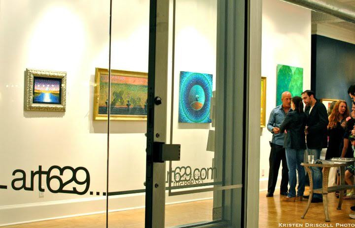 View inside an art gallery called art629 with people gathered, talking and looking at artwork.
