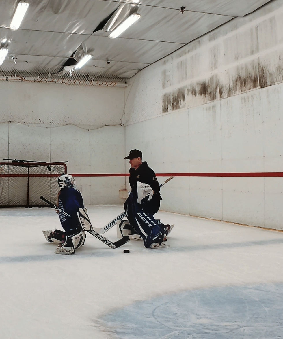 Goalie in dark gear kneeling on ice with coach in indoor rink