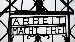 Wrought iron gate with the text "ARBEIT MACHT FREI" in bold letters. The background is cloudy and gray, conveying a somber mood.