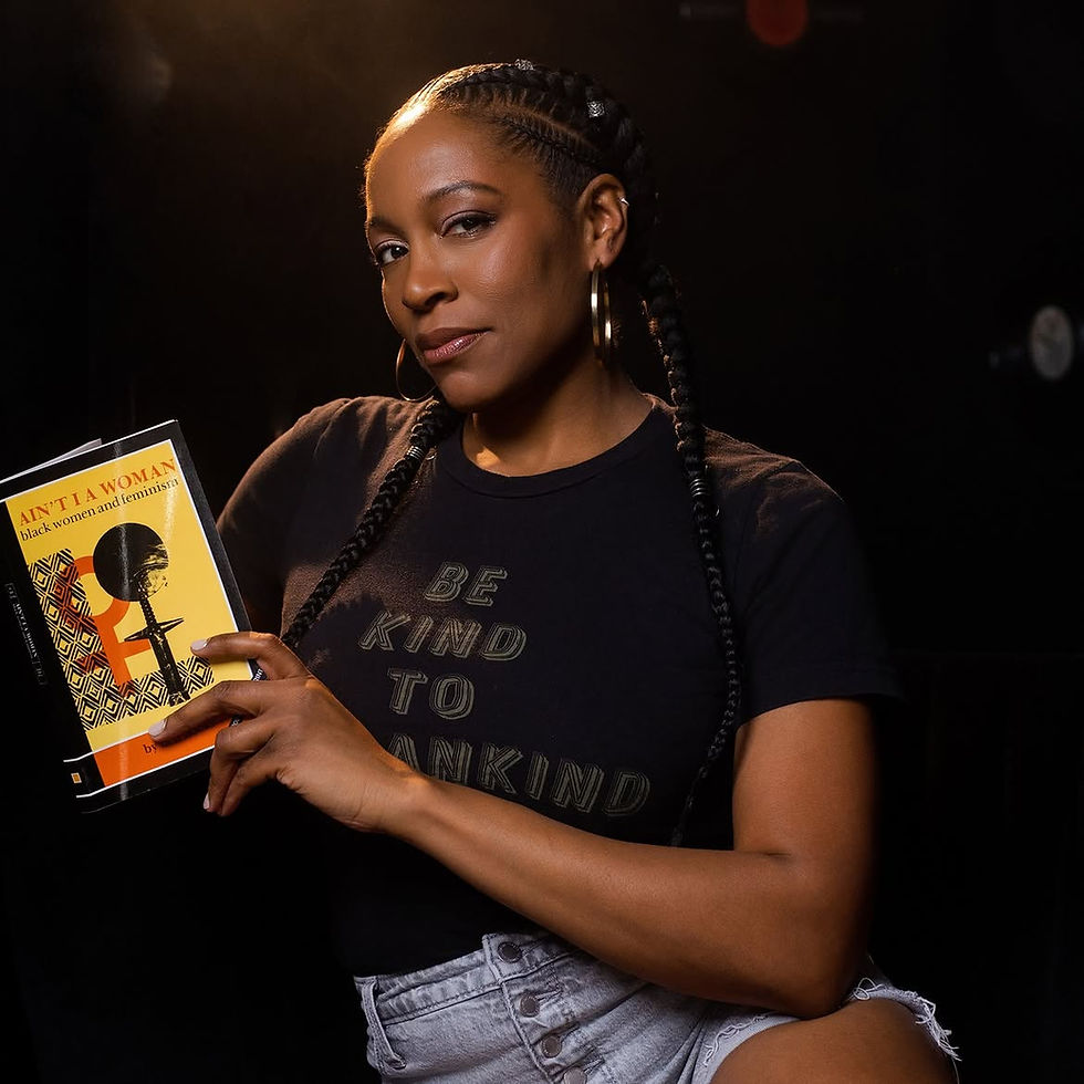 Esther Jade holds book Ain’t I a Woman; black shirt says Be Kind to Womankind against dark studio backdrop.