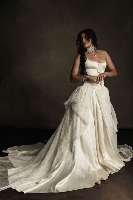 Rowyn showcases a meticulously fitted bodice, sculpting the body with precision and a soft satin finish. Thin, delicate straps highlight the décolletage and a silk chiffon bow at the neckline add a playful touch to its modern romantic design. Rowyn’s drop waistline enhances the figure, spilling into a voluminous bustled organza overskirt over cascading satin train to create a gorgeous balance of soft textures and romance.
