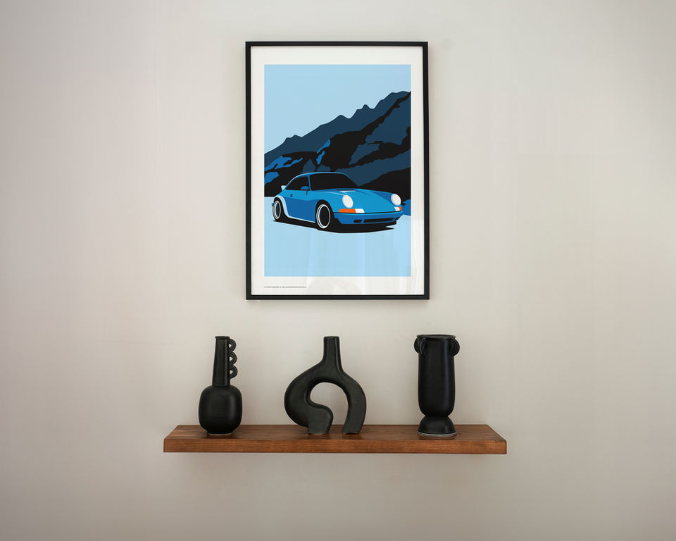 A framed art print of a blue Porsche 930 driving through a mountainous landscape hangs on a light gray wall. The illustration features dark blue mountains in the background and a bright blue sky. Below the artwork, a wooden floating shelf holds three modern black ceramic vases with abstract and sculptural shapes. The overall decor is minimalist and contemporary.