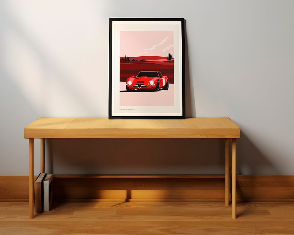 A framed art print of a red Alfa Romeo giulia TZ2 on a countryside road is resting on a minimalist wooden bench against a light gray wall. The illustration features a stylized red and pink landscape with distant hills and cypress trees. Two books are stored vertically in a small compartment beneath the bench. Natural light casts soft shadows across the floor and wall.