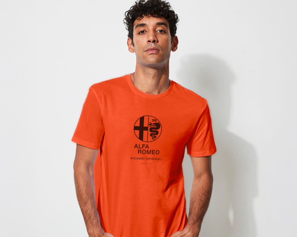 Cool young man with hands in pocket wearing an orange t-shirt featuring a screen-printed black Alfa Romeo logo and text reading ‘Ricambi Originali, Made in Italy’