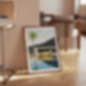 A framed modern art print of a house, palm tree, and yellow E-type Jaguar by a reflective pool is propped against a wall in a stylish, sunlit living room with wooden furniture.
