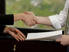 Two people shaking hands, one holding a document. Background shows a blurred office setting. The mood is professional and formal.