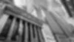 Black and white image of tall buildings with columns, adorned with American flags. A cloudy sky forms the backdrop, adding a dramatic effect.