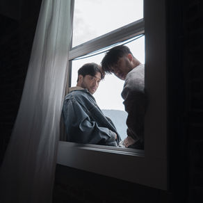 Mak Ro sitting together in an open window, looking toward the camera in a moody portrait framed by curtains and soft natural light.
