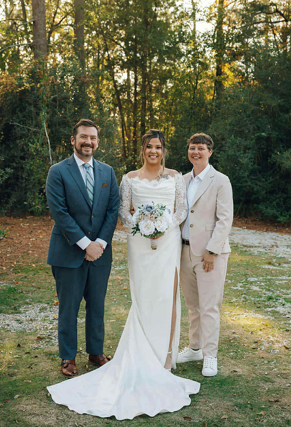 Three people smiling outdoors. A bride in a white gown holding flowers, flanked by two people in suits. Trees in the background.