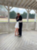 A couple embraces in a wooden gazebo with lattice walls. The woman wears a white dress, the man a suit. Trees and a white fence are visible.