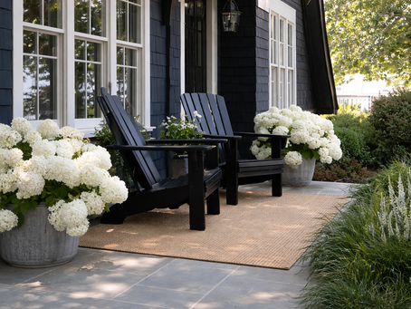 Black chairs on a porch with white hydrangeas in metal pots, against a dark blue house. Sunny greenery surrounds, creating a peaceful vibe.