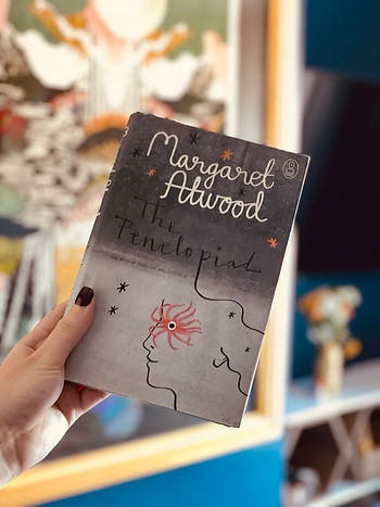 A woman's hand holding The Penelopiad book. The book cover shows Penelope surrounded by stars.