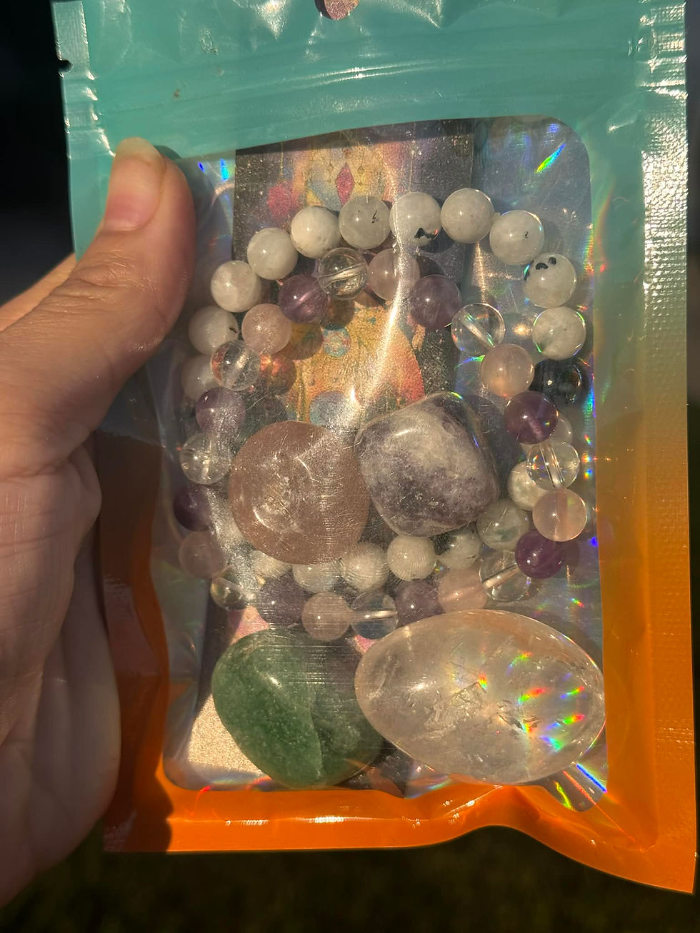 A hand holding a package of crystal beads, including amethyst, rose quartz, and clear quartz, with a rainbow reflection in the packaging.