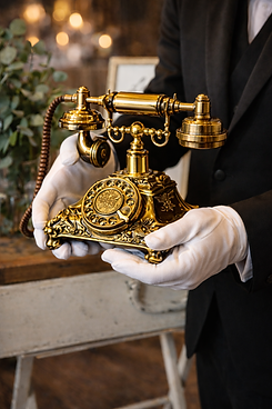 White-gloved attendant presenting an oversized antique gold audio guestbook telephone, with ornate rotary dial and handset, in a warm, elegant setting with soft bokeh lights and greenery in the background.
