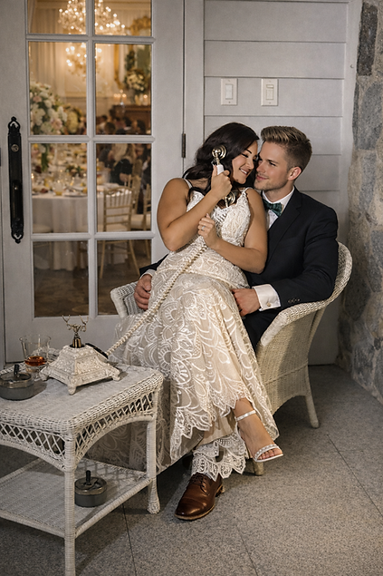 Elegant newlywed couple seated on a white wicker chair outside French doors, bride in lace gown holding a vintage audio guestbook phone while a softly lit wedding reception is visible through the glass panels behind them.
