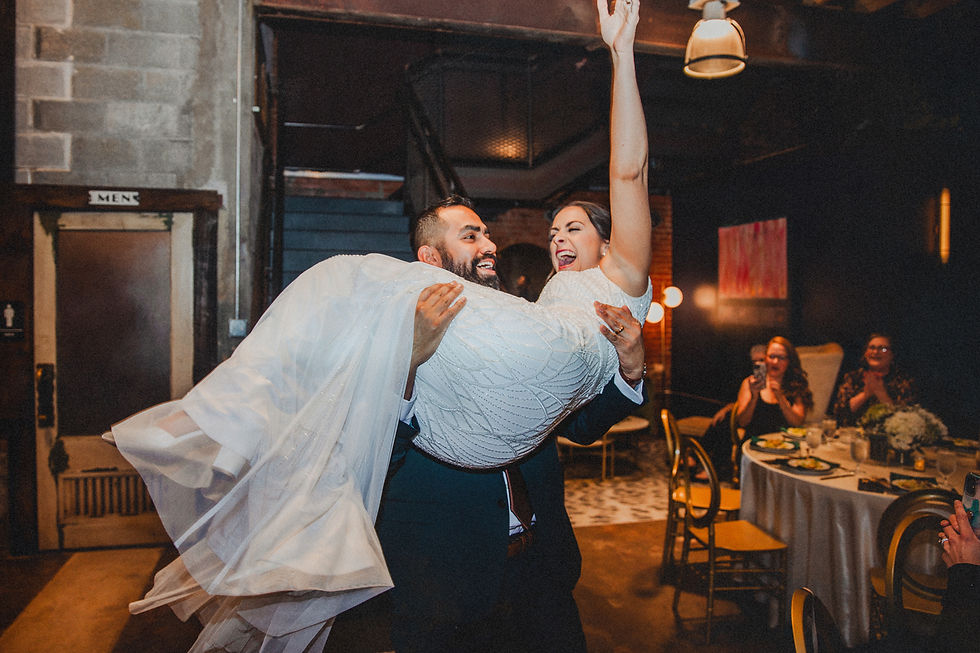 Groom lifts smiling bride in white gown in festive room. Guests clapping, visible "Men" sign, warm lighting, and joyful ambiance.