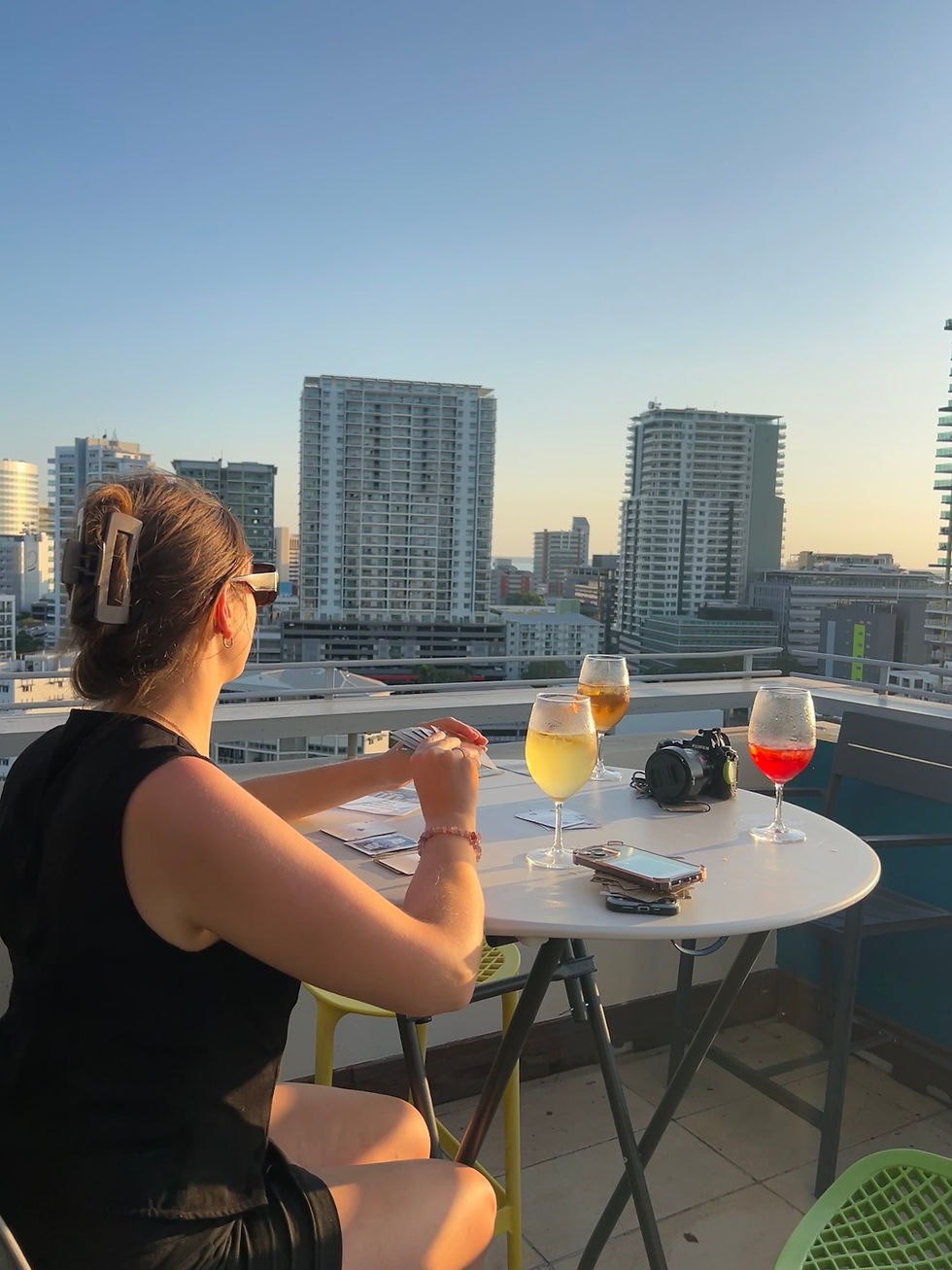 Woman sits at rooftop table with drinks, camera, and phone, overlooking city skyline at sunset. Calm and relaxed atmosphere.