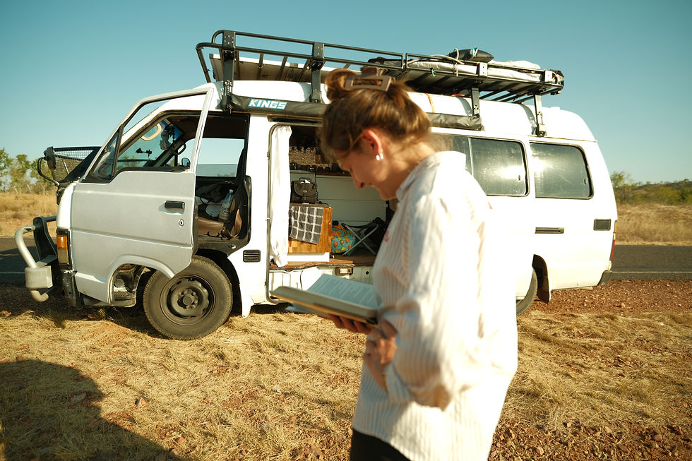 Woman reading outside a white van on a sunny day, parked on a dry, grassy roadside. Open van door shows packed interior. Calm mood.