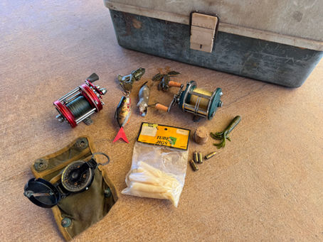 Fishing gear on a surface includes reels, lures, a compass, and an old tackle box. Pack of "Tube Jigs" and cartridge shells visible.