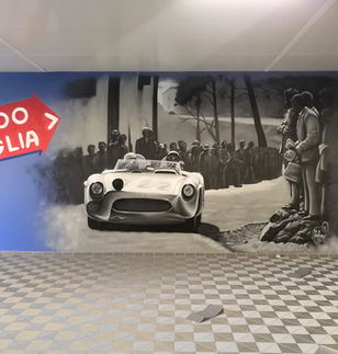 Une fresque murale de style street art illustre la voiture de course de Fangio filant sur les routes sinueuses de la 1000 Miglia. Les couleurs grises et les traits dynamiques évoquent la vitesse et l’adrénaline de cette célèbre épreuve italienne. En arrière-plan, le public présent et ce jour là pour assister à ce moment historique du sport automobile.