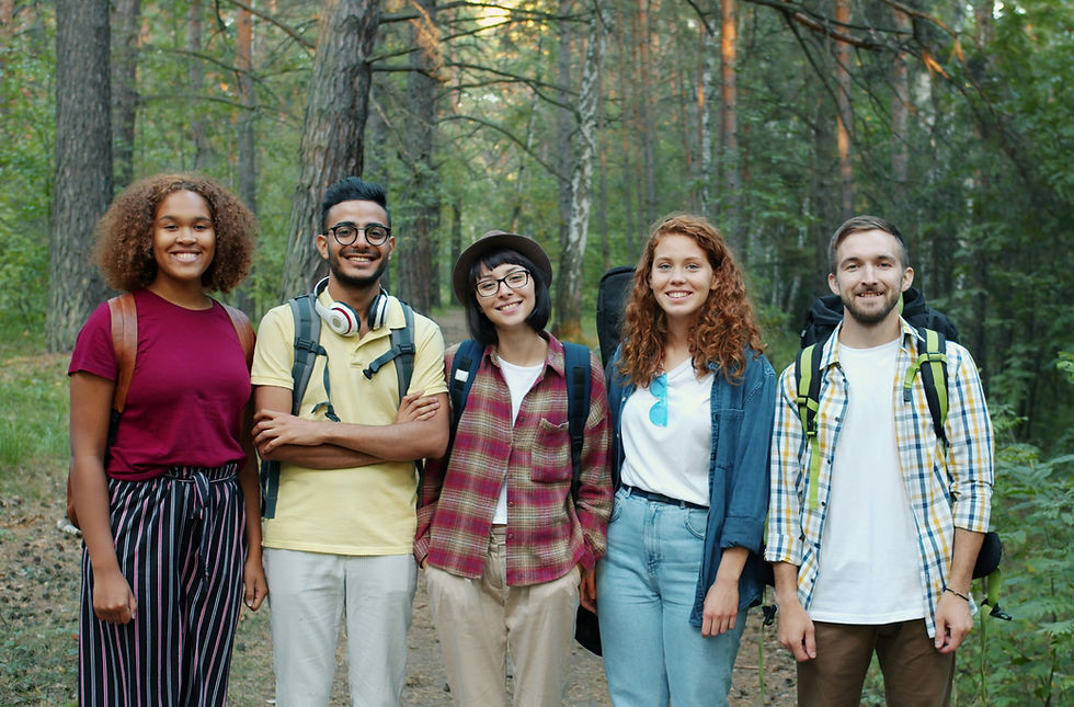 Five smiling hikers with backpacks pose on a forest trail among tall pine trees.