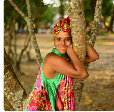 Black Woman therapist in colorful dress and headwrap leans on a tree, smiling. Background features a forest setting with soft light.