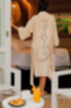 Woman in beige robe with moon phases enters bedroom. Coffee, juice, and croissant on table. Rattan lamp and greenery outside. Calm mood.