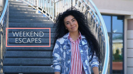 Woman with long curly hair standing on an outdoor staircase, with the Weekend Escapes travel series title displayed beside her.