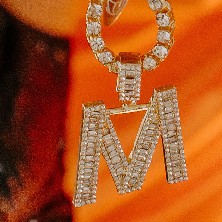 Close-up of a diamond-studded M pendant on a gold chain, hanging against a bright orange background.