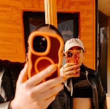 Two people take photos with phones in bold cases, standing against an orange wall. One wears a black jacket and cap with text.