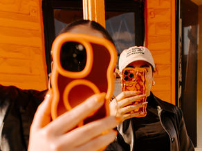 Two people take photos with phones in bold cases, standing against an orange wall. One wears a black jacket and cap with text.