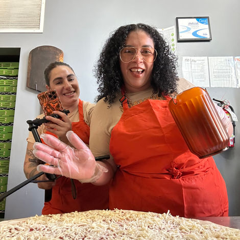 Two women in orange aprons with a large cheese pizza in front of them