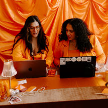 Two women in orange outfits work on laptops at a table with lamps, against draped orange satin, focused and collaborative.