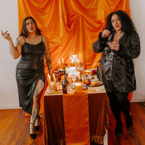 Two women in black sequined outfits pose by an orange draped table of candles, fruit, and drinks in a warm party scene