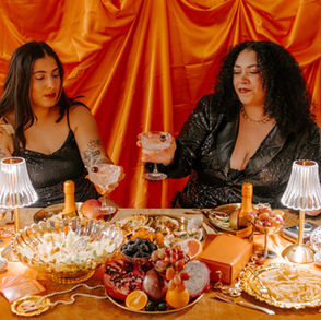 Two women in black seqence dresses with coup drinking glasses infront of table with snacks, wine, candles, lamps and more