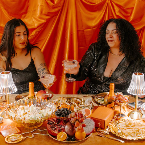 Two women in black seqence dresses with coup drinking glasses infront of table with snacks, wine, candles, lamps and more
