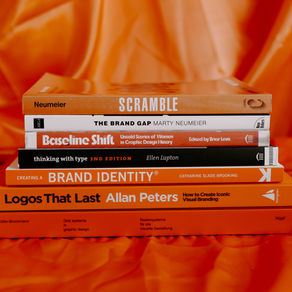 Stack of design books on orange fabric, with titles like SCRAMBLE, thinking with type, and Logos That Last visible.