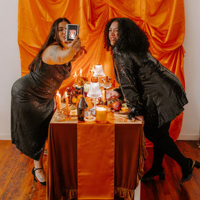 Two women in black sequin outfits pose for a selfie at a table with candles and fruit, against an orange draped backdrop, smiling.