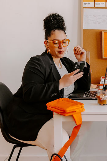 Woman with orange glasses checks phone at desk beside laptop, orange fabric, and corkboard with Pantone notes.