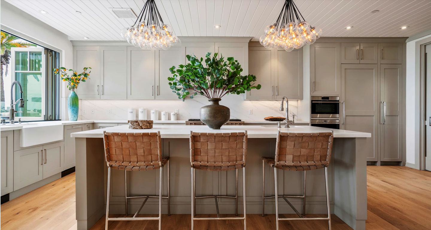 Warm light grey kitchen cabinets resemble agreeable grey, a versatile grey tone that pairs easily with other warmer tones throughout your space. This beach style kitchen features floor to ceiling cabinetry including customized areas built from the coordinating prefinished trim and panels. Warm wood floors, white board ceilings, and a large picture window create a welcoming atmosphere to sit at the matching kitchen island.