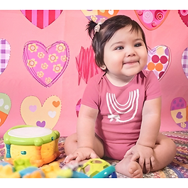 Lächelndes Baby in einem altrosa Body mit einem weißen, abstrakten Regenbogen-Motiv vor einem Hintergrund mit bunten Herzen