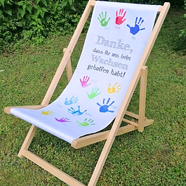Foto eines klappbaren Holz-Liegestuhls auf einer Rasenfläche. Die weiße Stoffbespannung ist mit vielen bunten Kinder-Handabdrücken, dazugehörigen Namen und dem grauen Schriftzug 'Danke, dass ihr uns beim Wachsen geholfen habt!' bedruckt.