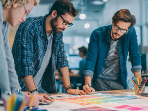 Three people in casual clothes collaborate on a colorful design project in a modern office. Mood is focused.