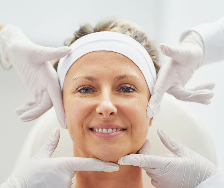 Eine junge Frau ist glücklich nach ihrer Gesichtspflege im Pro-Kosmetik-Studio.  Ihr Gesicht leuchtet vor Zufriedenheit über die erfolgreiche Hautverbesserung.  Ihre strahlende Haut wird liebevoll von den Händen zweier Kosmetikerinnen mit  weißen Handschuhen präsentiert.