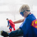 Young boy in superhero costume playing with Superman toy by the window.