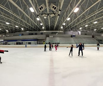 A view of Mountain View Ice Arena taken from the hockey benches, looking across center ice to the stands. Several skaters pass by, with a skater receiving a lesson on center ice.