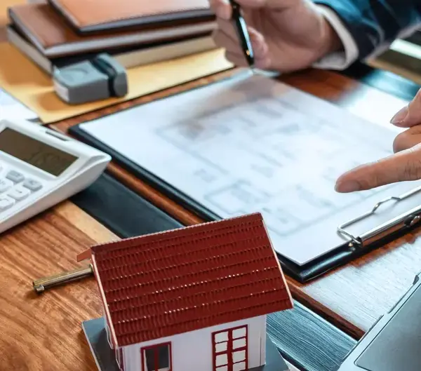 Person reviewing house plans on a tablet with a calculator and a small model home on the desk.