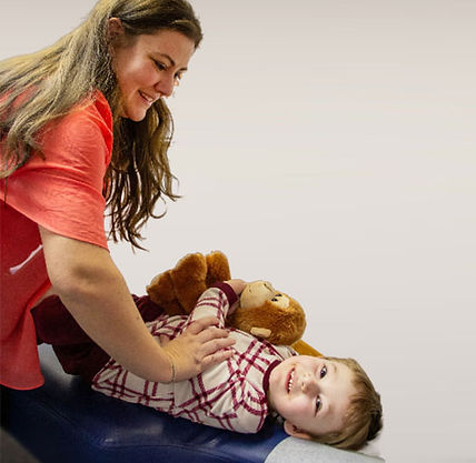 Smiling woman in coral shirt helps a smiling child lie on a blue table beside a teddy bear against a plain wall.
